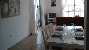Un comedor con una mesa preparada para cuatro personas, un televisor al fondo y un cuadro en la pared.
