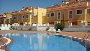 Una piscina con borde de azulejos, rodeada por un muro blanco bajo y una fila de edificios amarillos con tejados rojos.
