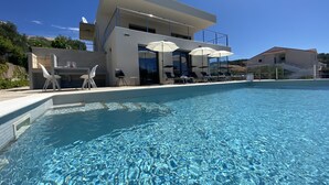 A modern poolside area with a clear pool, lounge chairs, umbrellas, and a dining setup. - Modern Villa with Pool & Sea View-40m to the Beach (Lun)
