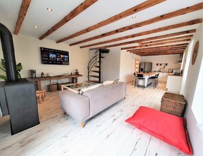 A modern living room with a sofa, a red cushion, a fireplace, wooden beams, a TV, and a kitchen area.
