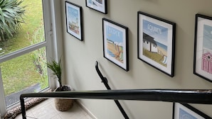 Un mur orné de plusieurs tableaux encadrés représentant des paysages côtiers, un escalier et une vue sur la verdure à l’extérieur.