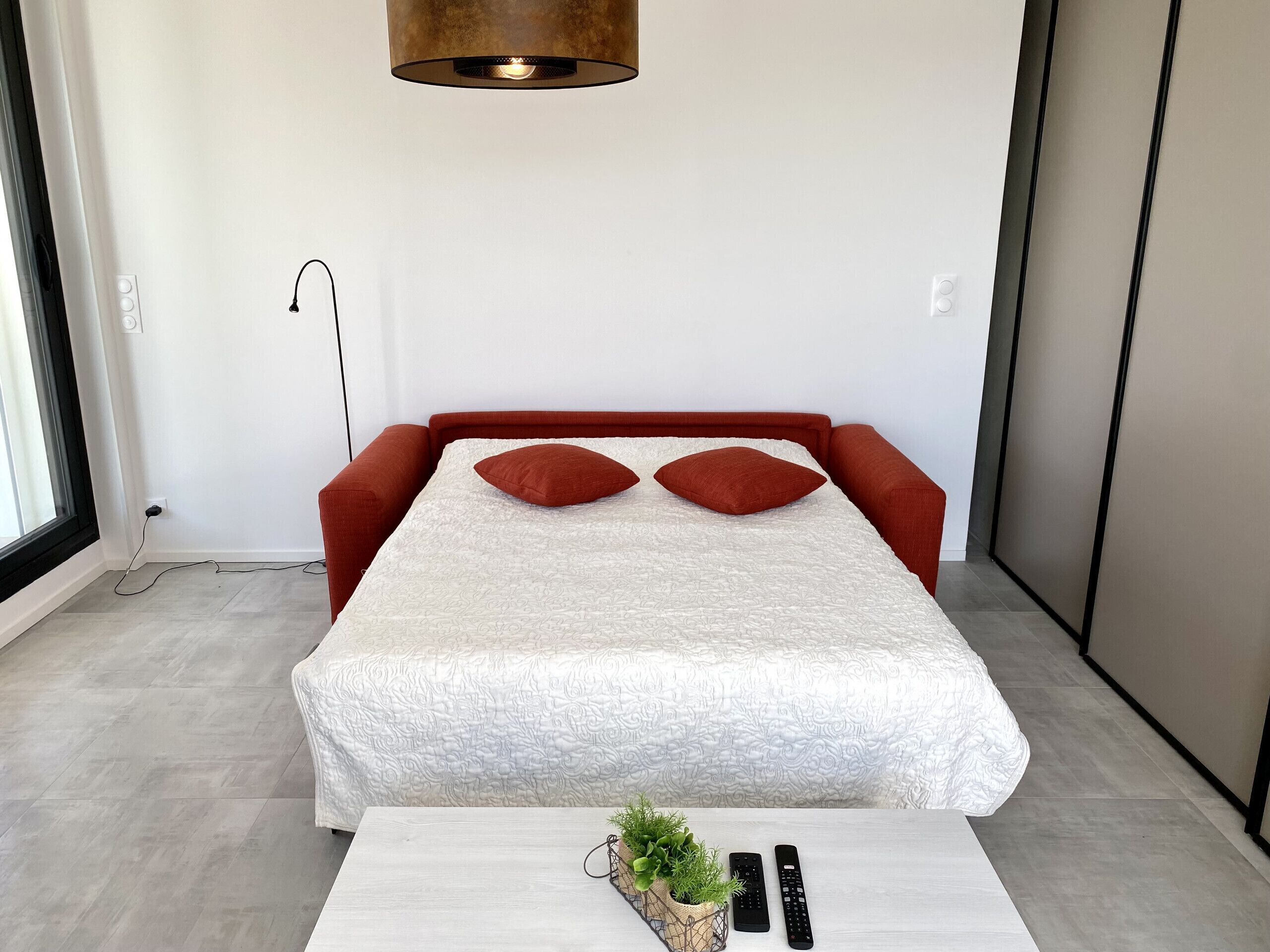 Ein modernes Schlafzimmer mit einem Bett, zwei Kissen, einem Nachttisch mit einer Pflanze und zwei Fernbedienungen.
