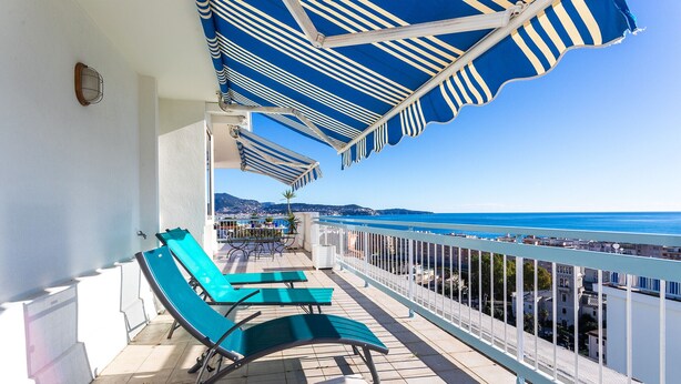 Un balcon agrémenté de parasols à rayures bleues, de fauteuils de jardin verts et offrant une vue sur la mer.