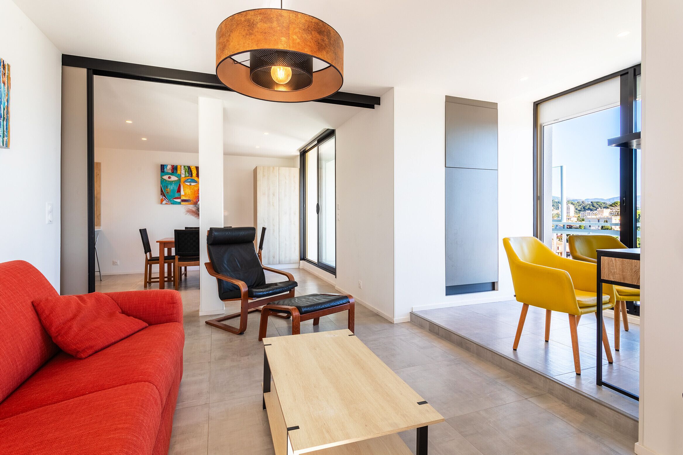 Un salon moderne avec un canapé rouge, une table basse en bois et des chaises jaunes. On y trouve également une chaise noire avec une structure en bois et un siège en cuir noir. Une grande fenêtre offre une vue sur la ville.