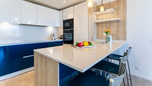 Une cuisine moderne avec des meubles blancs, un îlot central agrémenté d’un panier de fruits et un mur d’accent en bois.