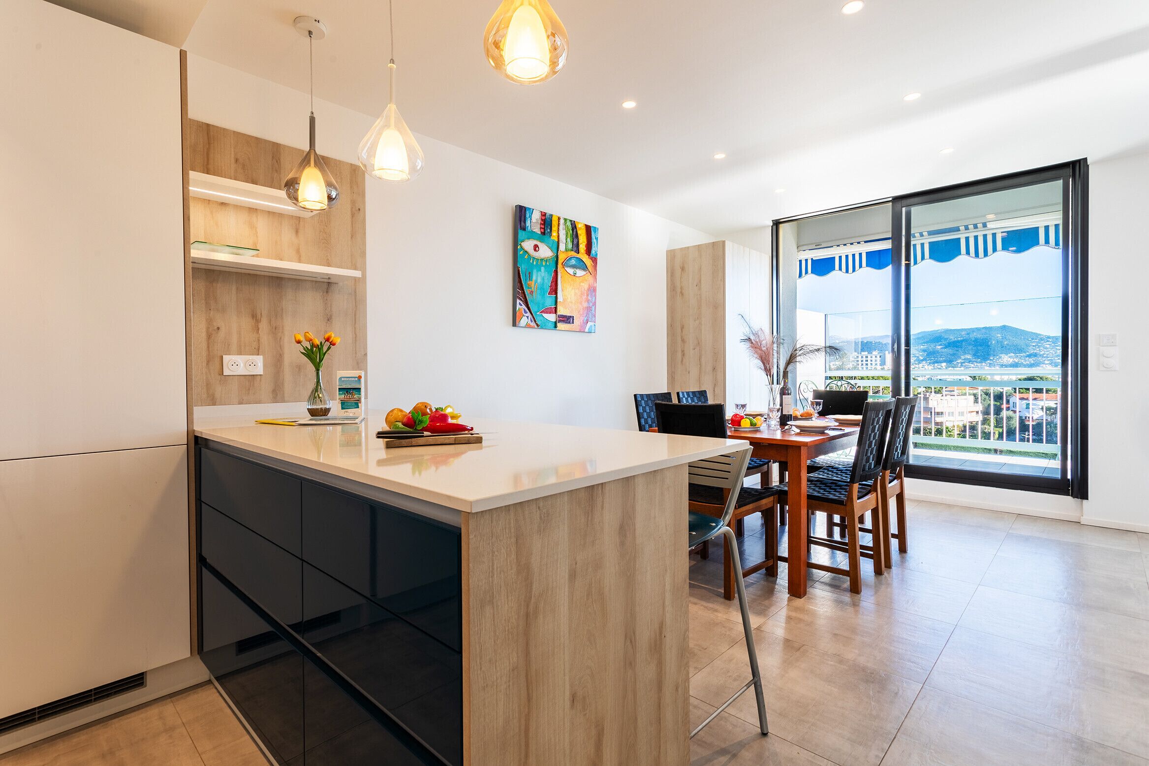 A modern kitchen with a central island, pendant lighting, and a dining area with a view of the city.