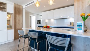 Une cuisine moderne avec des meubles blancs, un îlot central doté de tabourets de bar, un éclairage suspendu et un panneau d’accueil.