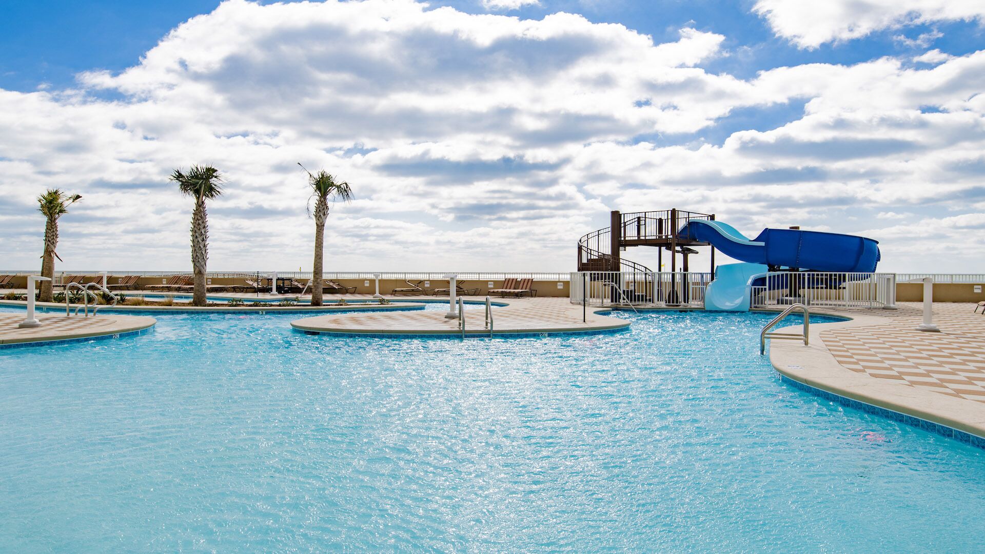 A swimming pool with a water slide, palm trees, and a clear sky.