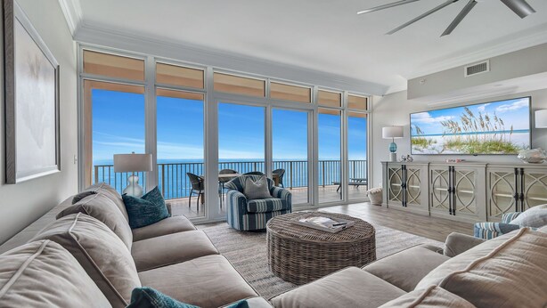A spacious living room with a large window overlooking the ocean, a comfortable sofa set, a wicker ottoman, and a television on a cabinet. - New Complex! Lazy River/Slide! Penthouse Level! (Orange Beach)