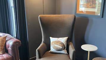A corner room with a dark-colored armchair, a small round table, a framed picture on the wall, and a window with curtains.