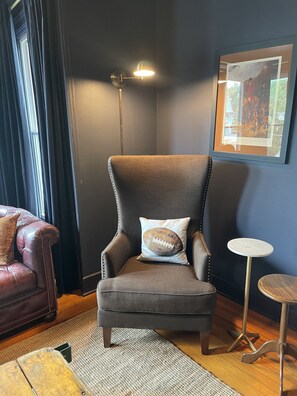 A corner room with a dark-colored armchair, a small round table, a framed picture on the wall, and a window with curtains.