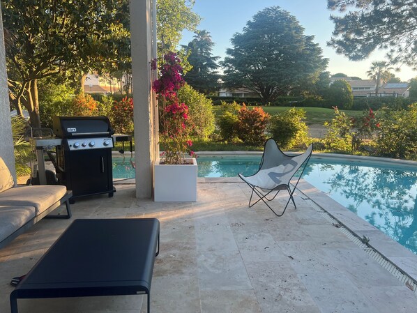 Zona de piscina con una parrilla, una silla de descanso y plantas en macetas.