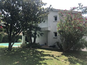 Un edificio de dos plantas con balcón, rodeado de vegetación y una piscina.