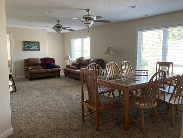 A spacious living room with a brown leather couch, a glass-top dining table with wooden chairs, two ceiling fans, and large windows with blinds.
