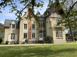 Une grande maison à étages, dotée d’une imposante cheminée en briques, de nombreuses fenêtres et d’une pelouse bien entretenue.