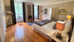 Ein geräumiges Schlafzimmer mit einem großen Bett, Holzboden, einem Sessel, einem Schreibtisch und einem Fenster mit Vorhängen.