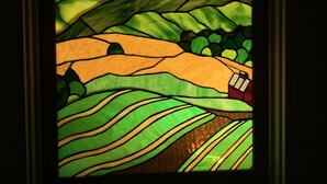 A framed stained glass window depicting a landscape with rolling hills and a house. - Vermont Gem Near Woodstock and Quechee (Woodstock)