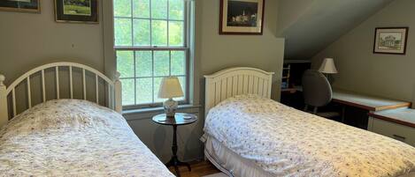 A bedroom with two single beds, a desk, a chair, a lamp, and framed pictures on the walls.