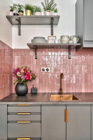 A kitchen with grey cabinets, a pink tiled backsplash, a sink, and a shelf with plants and dishes. - NEW! Eiffel Tour Splendid 2BR apt in Paris (Paris)