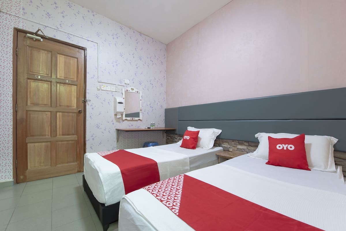 Une chambre d’hôtel avec deux lits, chacun recouvert d’une couvre-lit rouge et blanche, une porte en bois, un petit bureau avec un miroir et un mur orné d’un motif floral.