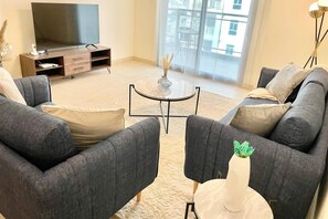 A modern living room with a sofa set, a coffee table, a flat-screen TV on a wooden stand, and a large window with a view of buildings.