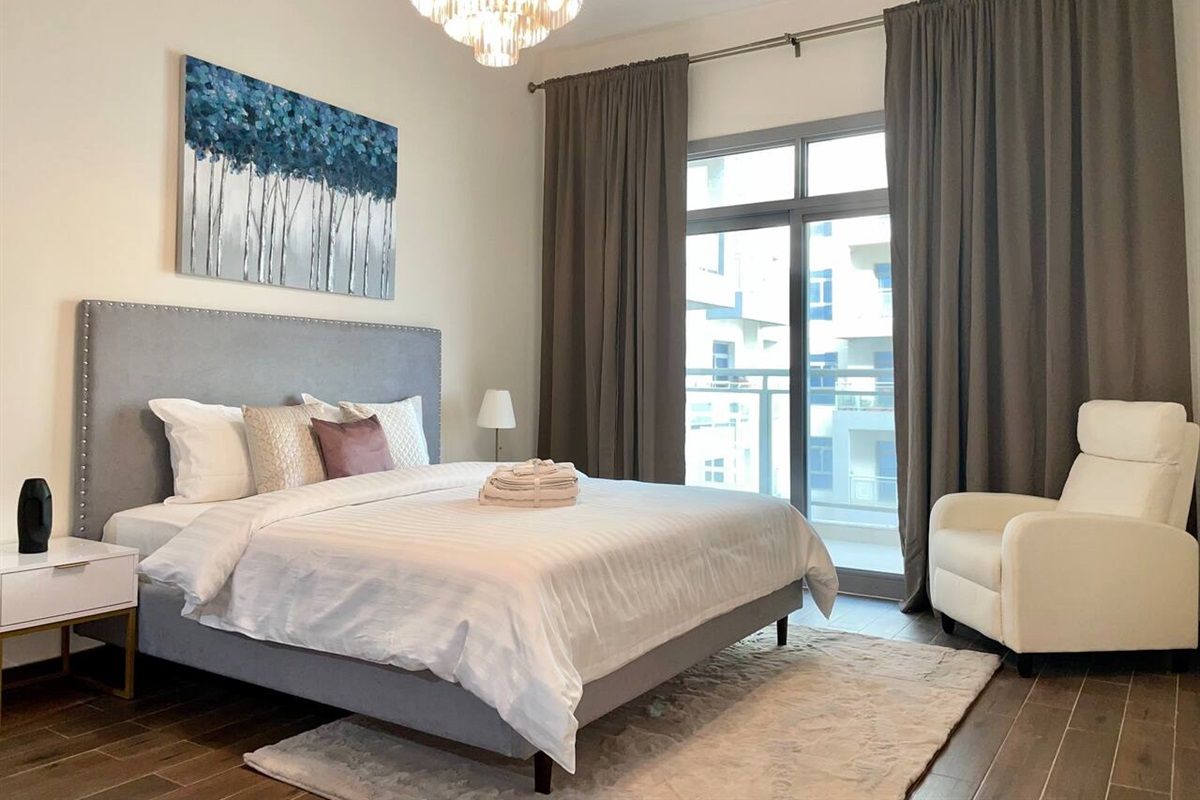 A bedroom with a large bed, a nightstand, a chair, a window with curtains, and a painting on the wall.