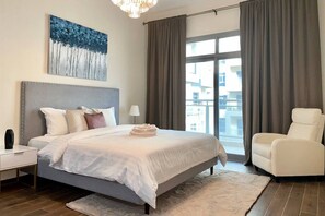A bedroom with a large bed, a nightstand, a chair, a window with curtains, and a painting on the wall. - Spacious 2BR with Direct Metro Access (Dubai)
