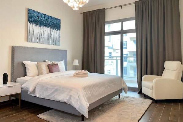 A bedroom with a large bed, a nightstand, a chair, a window with curtains, and a painting on the wall.