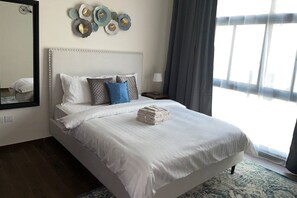 A neatly made bed with white linens and a stack of folded towels, a wall clock, and a window with curtains. - Spacious 2BR with Direct Metro Access (Dubai)