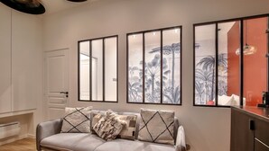 Una sala de estar moderna con un sofá gris, una puerta blanca y un ventanal con un mural tropical.