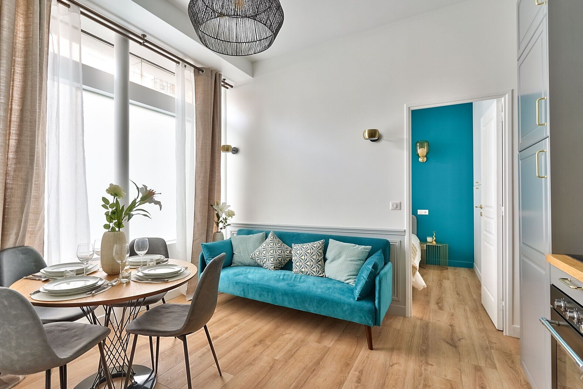 A modern living room with a teal sofa, a dining table set for four, and a kitchen visible through an open door.
