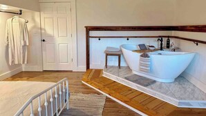 A bedroom with a large white bathtub, wooden flooring, a white bed, and a door. - King Apt Suite  w/ Soaker Tub & Mountain Views (Bryson City)
