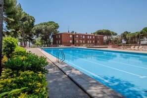 Piscina com água azul cristalina, rodeada por vegetação e um edifício ao fundo.
