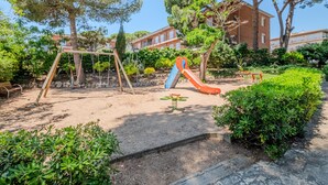 A playground with swings, a slide, and a seesaw in a landscaped area with trees and a building in the background.