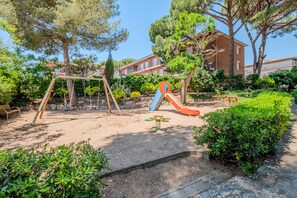 A playground with swings, a slide, and a seesaw in a landscaped area with trees and a building in the background.