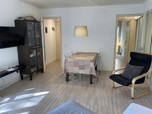 Un salon comprenant un téléviseur, un meuble, une table à manger avec des chaises, une lampe suspendue et un fauteuil avec un coussin.