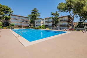Une piscine entourée d’une terrasse carrelée, elle-même bordée d’immeubles à plusieurs étages dotés de balcons, d’arbres, et sous un ciel dégagé.