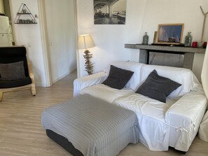 Un salon avec un canapé blanc, un fauteuil noir, une lampe, une petite plante et une cheminée ornée d’objets décoratifs.