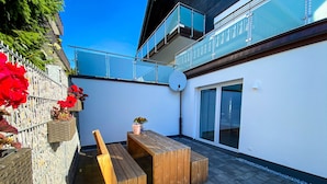 Un patio con un banco y una mesa de madera, una pared blanca y un balcón de cristal.