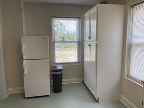 A compact kitchen with a white refrigerator, a trash bin, and a window with blinds. - Walk To The Beach At Beacon Hill. (Port St. Joe)