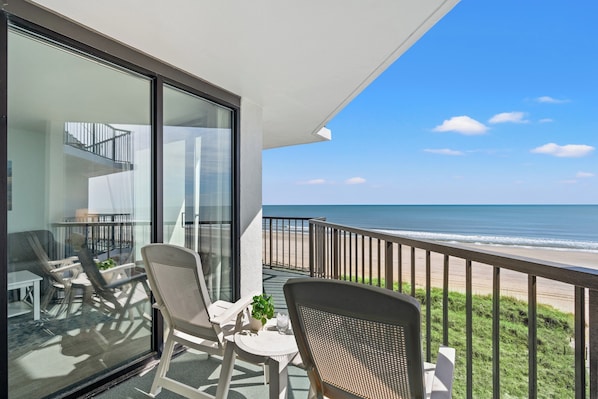 Uma varanda com vista para a praia, uma mesa com cadeiras e um vaso de planta.
