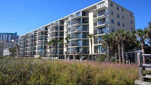 Un hôtel à plusieurs étages, doté de balcons, situé près d’une plage bordée de palmiers.