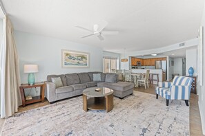 A living room with a sectional sofa, a coffee table, and a striped armchair. There is a kitchen area with wooden cabinets and a microwave.