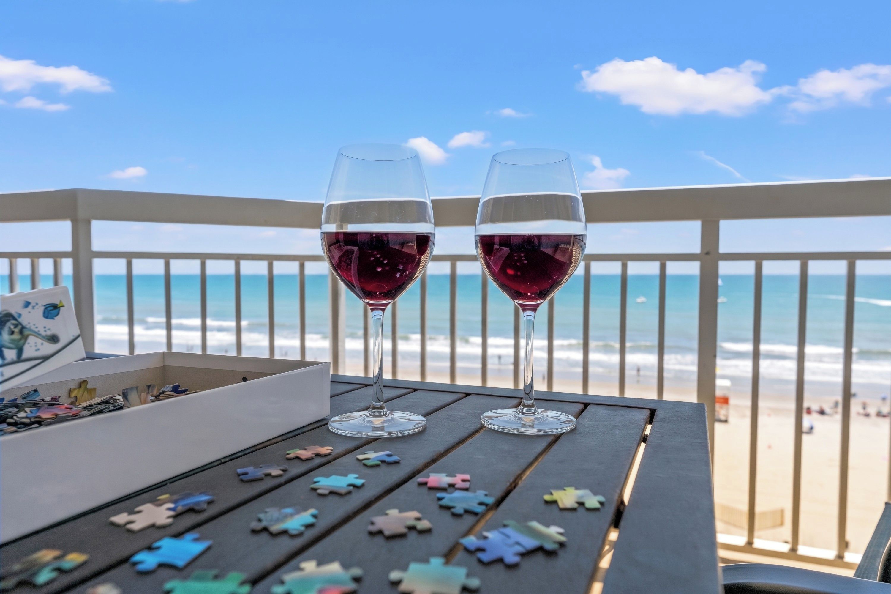 Two wine glasses on a table with puzzle pieces, overlooking a beach with a railing.