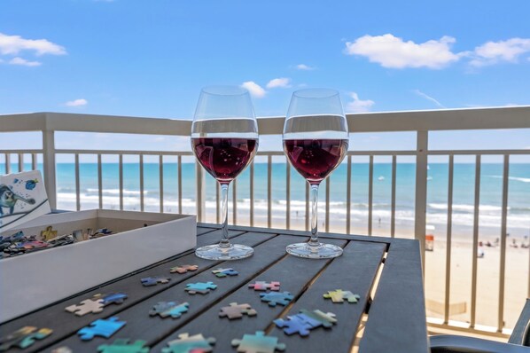Two wine glasses on a table with puzzle pieces, overlooking a beach with a railing.