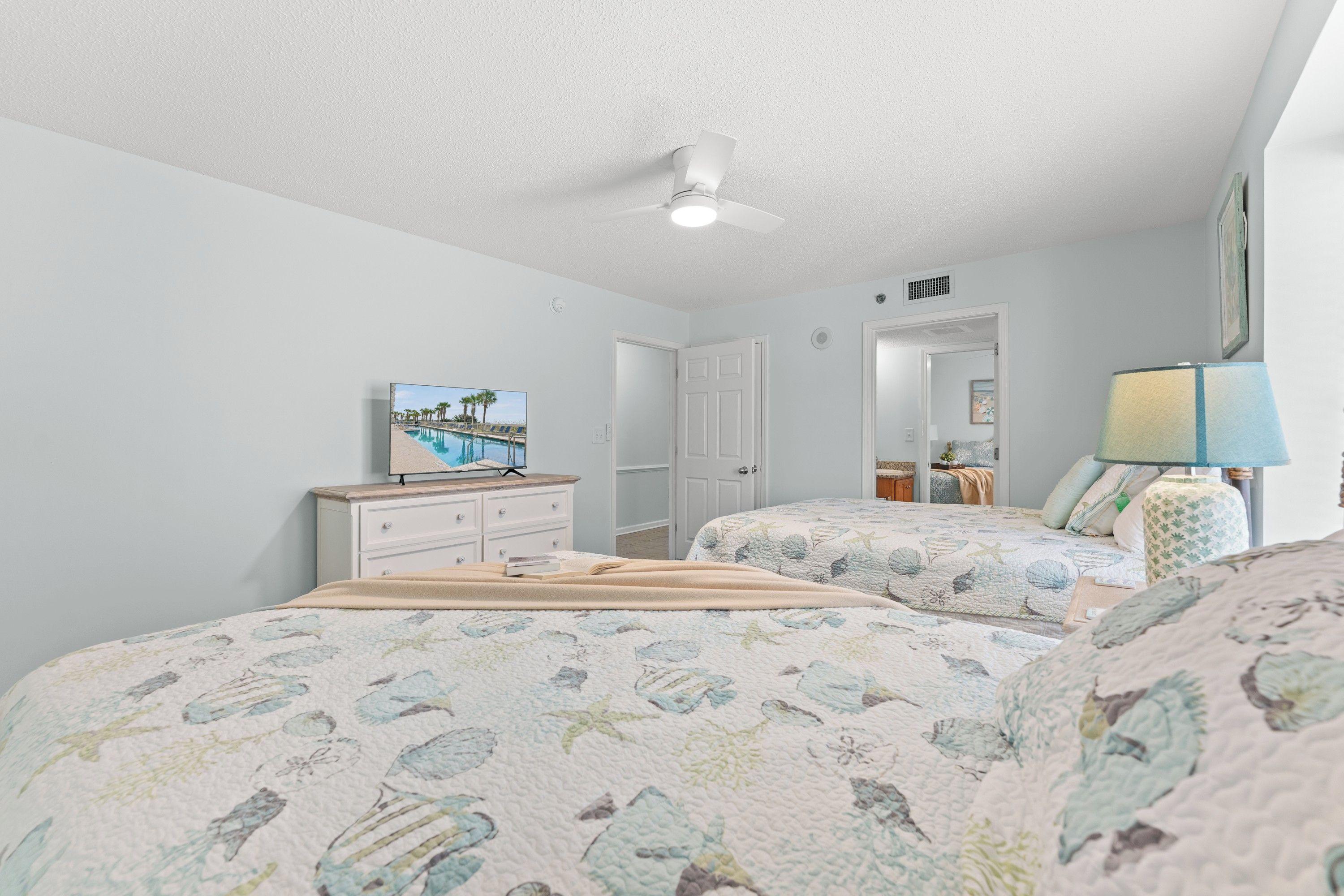 Une chambre avec deux lits, un ventilateur de plafond, un tableau et une lampe.