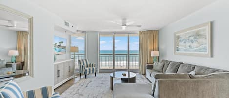 A spacious living room with a large window overlooking the beach, a ceiling fan, and a painting on the wall.