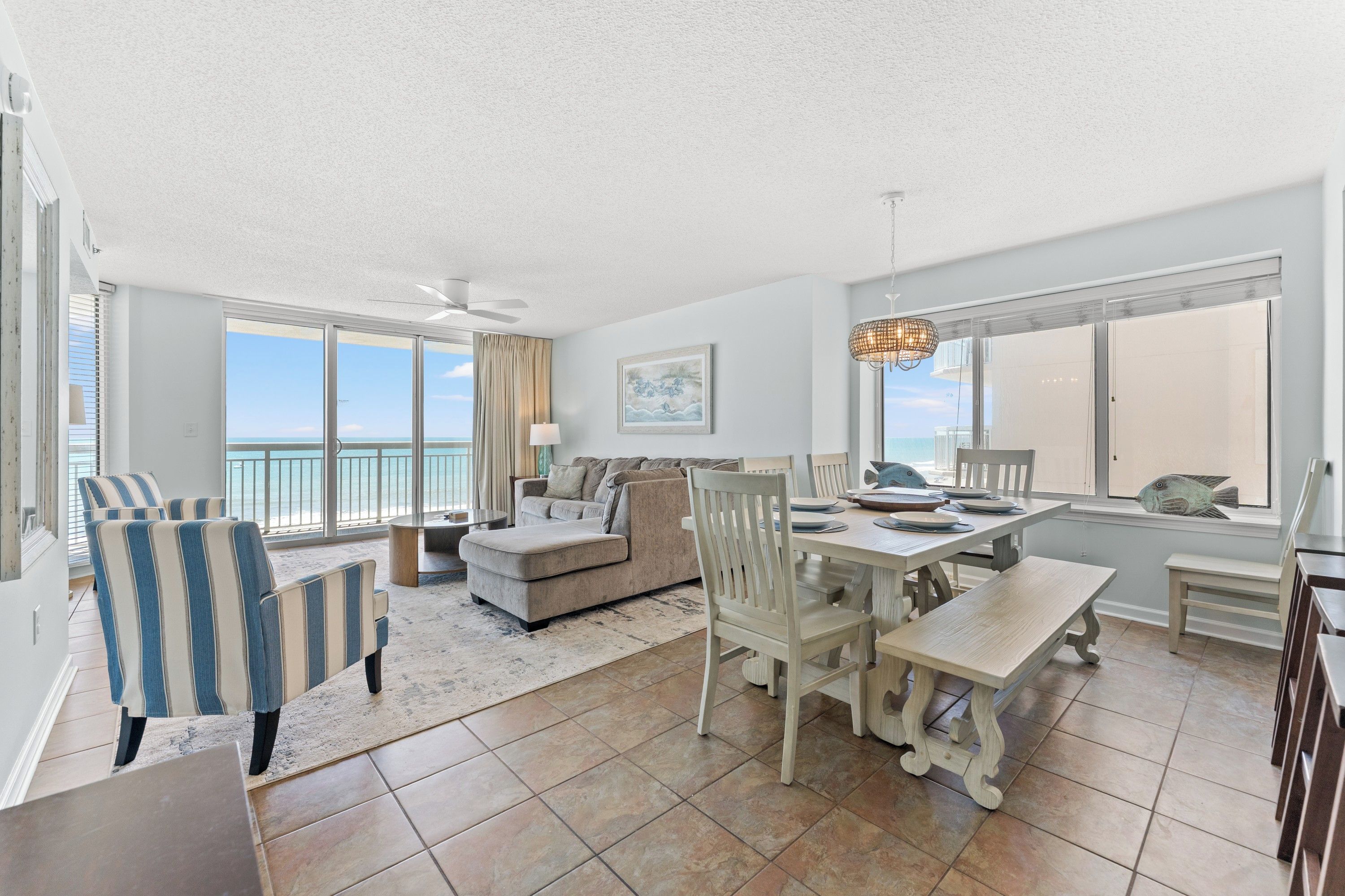 A spacious living area with a large window overlooking the ocean, a dining table set for four, and a ceiling fan.