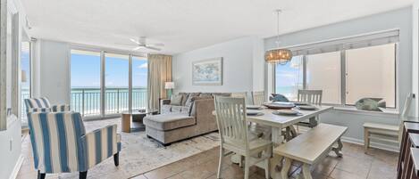 A spacious living area with a large window overlooking the ocean, a dining table set for four, and a ceiling fan.