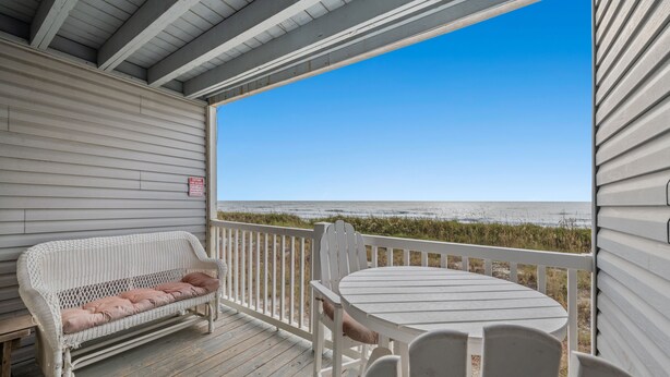 A wooden deck with a white bench and table, overlooking a beach. - Nautical Watch 207-1BR/1BA Oceanfront condo in Windy Hill (North Myrtle Beach)